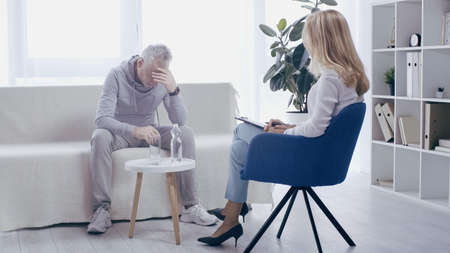 depressed mature sportsman and blonde psychologist having conversation in consulting roomの写真素材