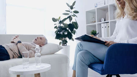 psychotherapist writing on clipboard near blurred middle aged man with clenched hands lying on couch