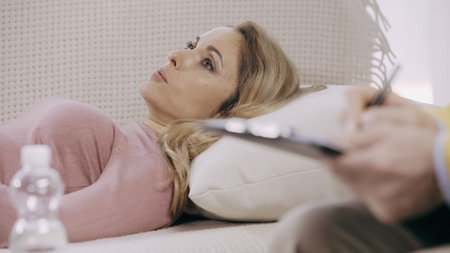 blurred psychotherapist writing on clipboard near blonde patient lying on couch in consulting room