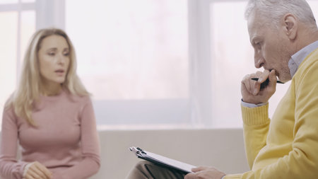 thoughtful middle aged psychotherapist holding pen near clipboard and listening blurred patient in consulting roomの写真素材