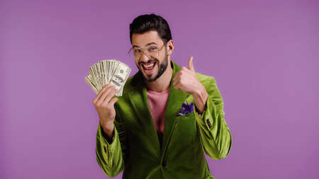 happy man in green jacket and glasses holding dollars and showing thumb up isolated on purpleの写真素材