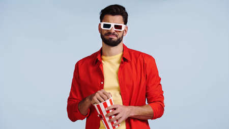 dissatisfied man in 3d glasses watching movie and holding popcorn bucket isolated on blue