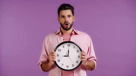 shocked young man holding clock isolated on purpleの写真素材