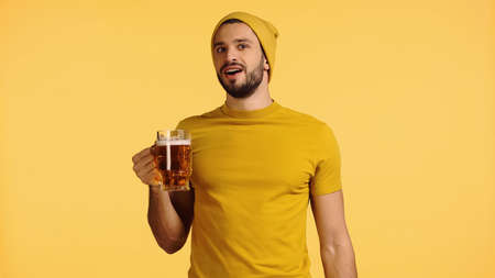 amazed man in beanie hat and t-shirt holding glass mug with beer isolated on yellowの写真素材