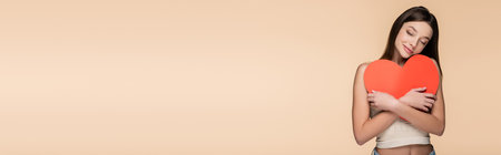 joyful woman with closed eyes hugging red paper heart isolated on beige, bannerの写真素材