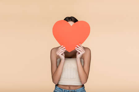 young woman in crop top with bare shoulders covering face with red paper heart isolated on beigeの写真素材