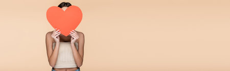 young woman in crop top with bare shoulders covering face with red paper heart isolated on beige, bannerの写真素材