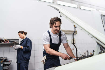 Positive mechanic working with wrench and car near blurred colleague in service