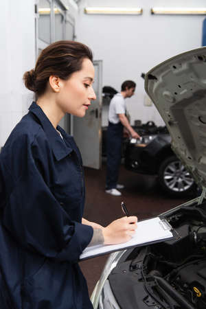 Side view of mechanic writing on clipboard near car with open hoodの写真素材