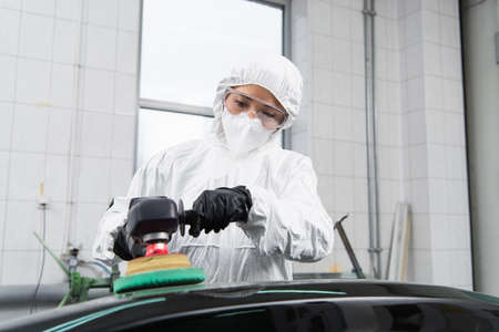 Workwoman in hazmat suit and protective mask polishing car in service