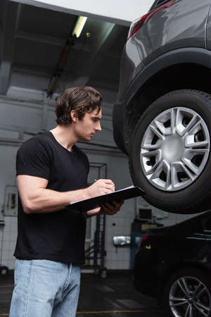 Side view of mechanic writing on clipboard near car wheel in garage