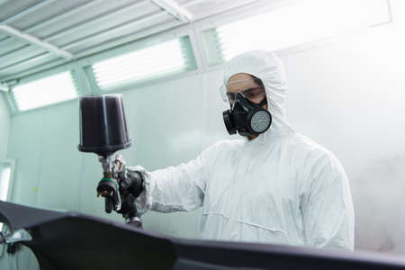 Workman in hazmat suit and respirator coloring blurred car part in service