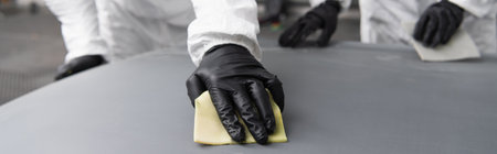 Cropped view of workman in hazmat suit and gloves using sandpaper on car hood in garage, banner