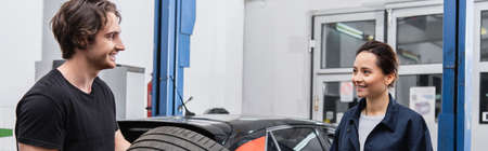 Smiling mechanic looking at colleague near tire and car in service, banner