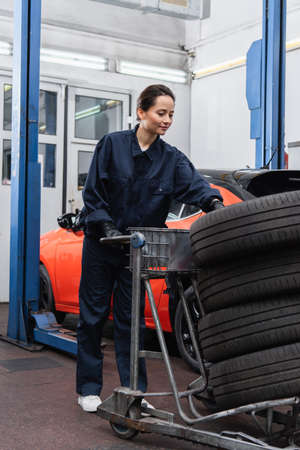 Workwoman in uniform standing near cart with tires and car in garage