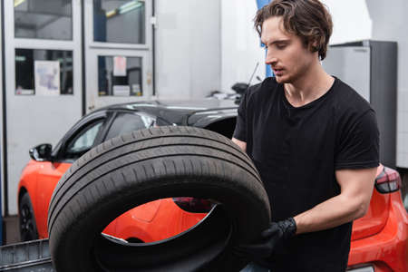 Young workman in gloves holding tire near blurred car in serviceの写真素材
