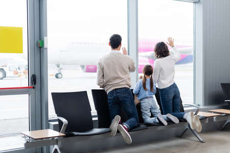 back view of family waving hands while looking at plane through window in airport