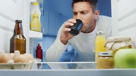 man drinking soda from metal can near fridge with beverages, eggs and bottles with sauces
