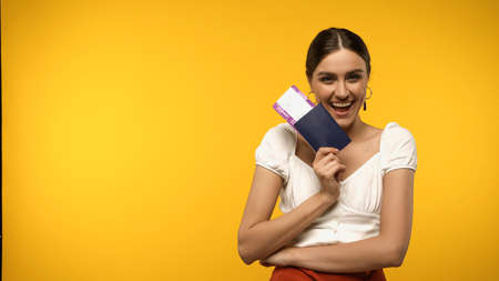 Happy woman holding passport and air ticket while looking at camera isolated on yellowの写真素材