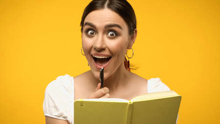 Excited woman in blouse holding pen near notebook isolated on yellow
