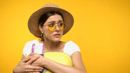 Scared woman in sunglasses holding baggage and passport isolated on yellowの写真素材