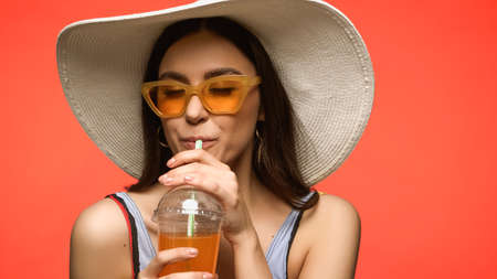 Pretty woman in sunglasses and swimsuit drinking cocktail isolated on coralの写真素材