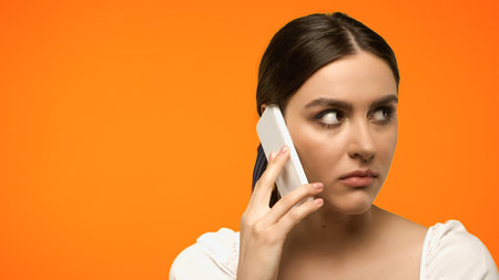 Brunette model looking away while talking on smartphone isolated on orange