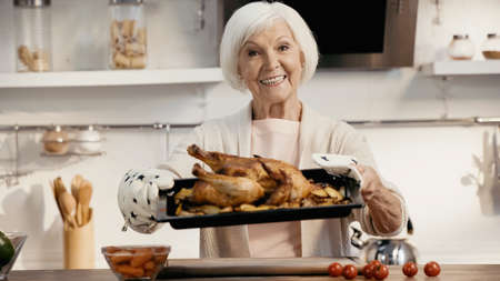 happy senior woman holding oven sheet with roasted turkey near fresh cherry tomatoes and carrots in kitchenの写真素材