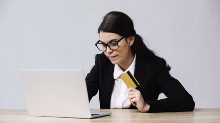 businesswoman in glasses holding credit card near laptop isolated on grey