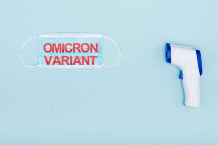 top view of medical mask with red omicron variant lettering near pyrometer on blueの写真素材