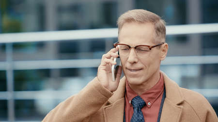 Smiling businessman in coat talking on mobile phone on urban street