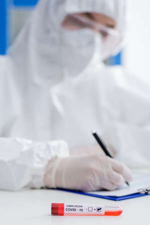 blurred scientist in hazmat suit writing on clipboard near positive covid-19 omicron variant test in...