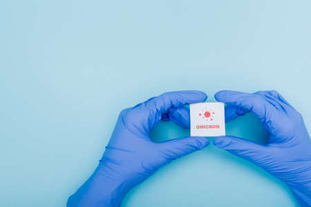 white card with red omicron lettering and bacteria icon in hands of doctor in latex gloves on blue, cropped viewの写真素材