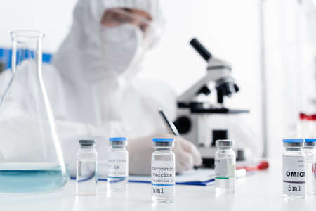 selective focus of vials with coronavirus vaccine near scientist writing near microscope on blurred...