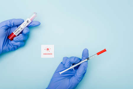 top view of doctor in latex gloves holding syringe and test tube near card with omicron lettering and bacteria on blueの写真素材
