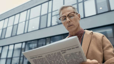 Low angle view of mature businessman in coat and eyeglasses reading news on urban street