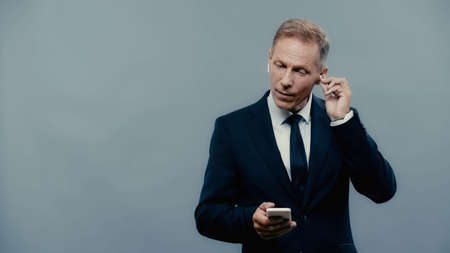 Mature businessman in formal wear using smartphone and earphones isolated on greyの写真素材