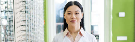 asian oculist in white coat looking at camera near blurred assortment of eyeglasses in optics salon,...