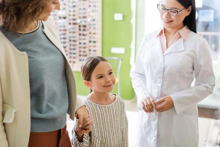 pleased mother and daughter holding hands near asian oculist in optics store