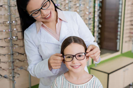 smiling asian oculist choosing eyeglasses for happy girl looking at camera in optics storeの写真素材