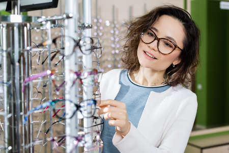 young positive woman choosing eyeglasses in optics salon on blurred foreground