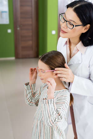 positive asian ophthalmologist in white coat trying eyeglasses on child in optics salon