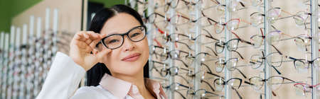 happy asian oculist looking at camera and touching eyeglasses in optics salon, banner