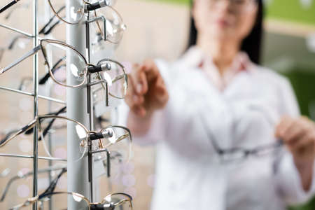 partial view of blurred oculist pointing at eyeglasses in optics shop