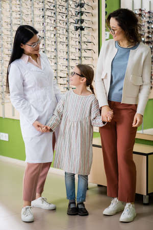 happy girl in eyeglasses holding hands with mother and asian oculist in optics shop
