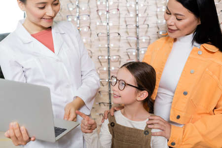 smiling girl in eyeglasses pointing at laptop near oculist and asian mother in optics salonの写真素材