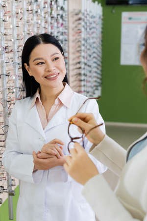 blurred woman choosing eyeglasses near smiling asian ophthalmologist in optics shop