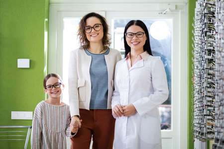 happy asian oculist and mother with daughter smiling at camera in optics shop