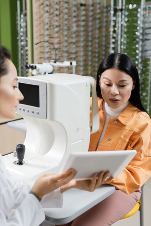 blurred oculist showing digital tablet to asian woman near vision screener in optics salon