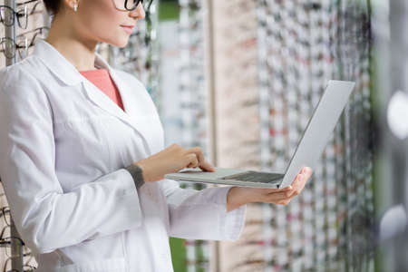cropped view of oculist in white coat using laptop in optics shop on blurred background